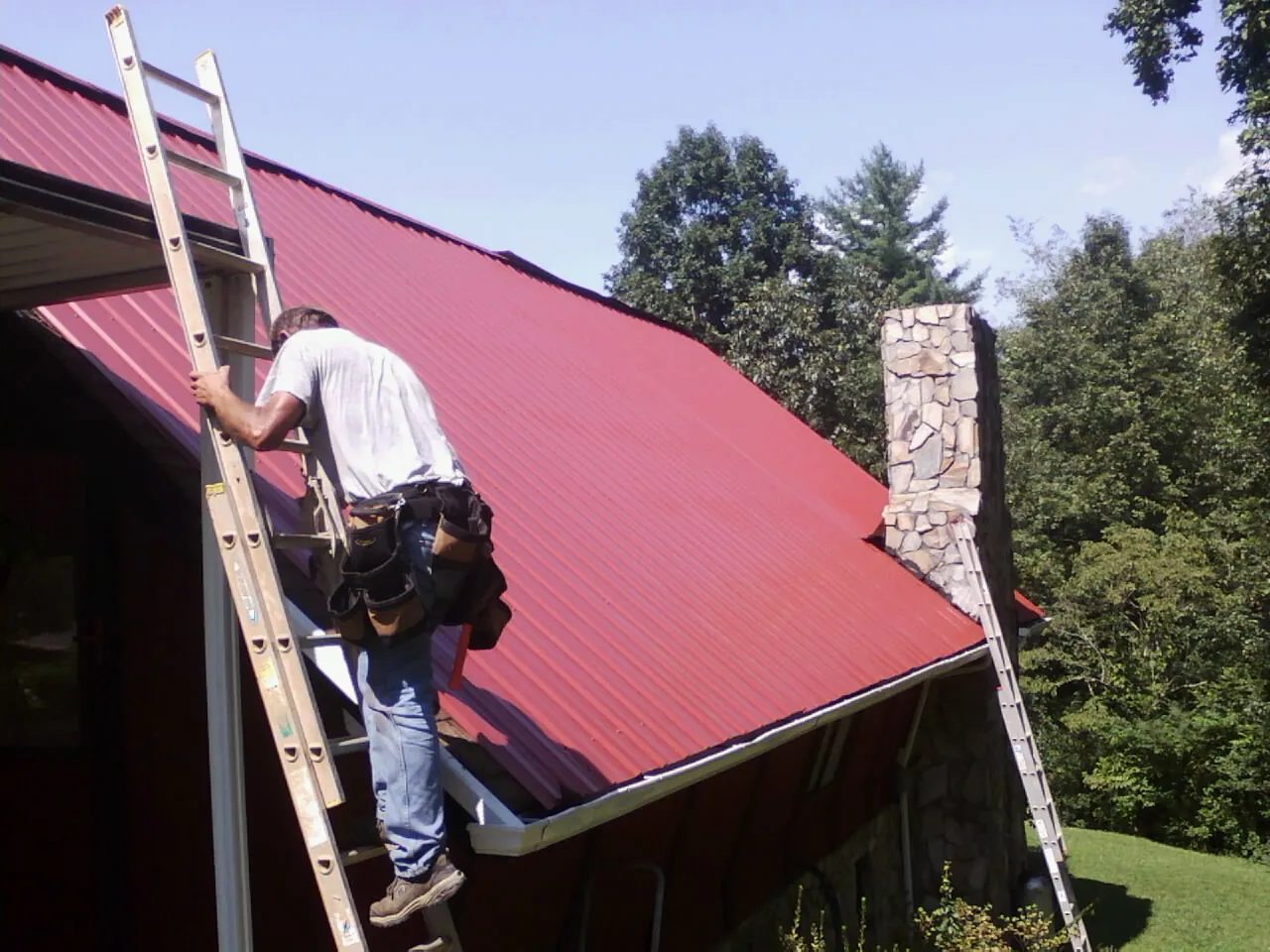 Roofer on a ladder beside a red metal roof and stone chimney — Soffit & Fascia Repair by Roofing Brothers in Englewood